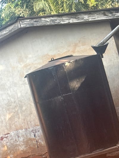 A dark, rectangular object dominates the view. It appears to be metallic, possibly a container or duct, with visible seams and a slightly uneven surface texture. A silver, angled pipe connects to its top. The structure is set against a light-colored wall, with a glimpse of green foliage visible on the left. The composition and focus suggest a shot taken indoors, looking upwards. The lighting creates highlights and shadows on the surfaces, enhancing their textures.
