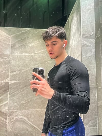 A young man with short, dark, messy hair and a light beard is taking a mirror selfie in a gym bathroom. He is wearing a black long-sleeved athletic shirt and blue shorts. He has white wireless earbuds in his ears and is holding a silver smartphone in his right hand. The bathroom has grey tiled walls and a dark, reflective surface behind him. The lighting is dim, with a green tint in the upper portion of the image. The location is the gym bathroom in Harmanli, Bulgaria.