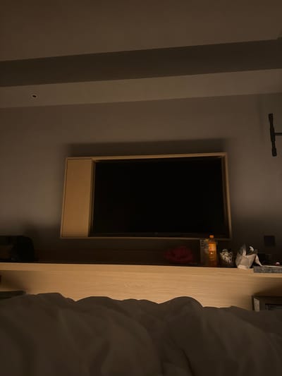 The image depicts an indoor scene, likely a bedroom, with a focus on a wall-mounted television. The television is framed in a light wood, and below it is a matching wood cabinet. To the right of the television, a tall, light wood closet or shelving unit is visible. This unit contains several items, including what appears to be bottles, a small patterned object, and a red loofah or sponge. A portion of a white, rumpled duvet or blanket is prominently featured in the foreground on the right side of the image, suggesting a bed is nearby.
The room appears to be dimly lit, with the television screen being a dark, blank rectangle, indicating it is either off or displaying a dark image. The lighting suggests it could be evening or nighttime. The wall to the left of the television is a plain, neutral color, with some shadows creating a sense of depth. Above the television, a dark industrial-looking fixture is mounted on the wall.
The overall mood is quiet and still, with no people present. The setting appears to be modern and minimalist. The context provided places the location in Chaoyang District, China. There is no visible text in the image.