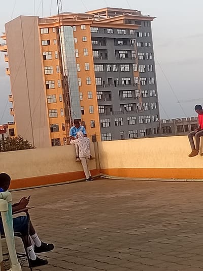 The scene depicts an urban rooftop in Karingani ward, Kenya, featuring three male individuals. The rooftop surface is paved with dark grey interlocking bricks laid in a herringbone pattern. A beige-colored parapet wall with an orange stripe along its base runs across the midground.
On the far left, a male is seated in a light-colored chair, looking down at a mobile phone held in his hands. He wears a blue top, dark shorts, white socks with a black logo, and black shoes. In the center, another male is perched atop the parapet wall, facing left, wearing a light-colored patterned hoodie, light-colored pants, and black shoes. On the far right, a third male is also perched on the parapet wall, facing right, dressed in a red shirt, dark pants, and light-colored shoes.
In the background, two prominent multi-story buildings are visible. The building to the left is predominantly beige/orange, featuring numerous windows and a tall vertical glass panel on its facade, with an antenna or mast-like structure extending from its roof. The building to the right is largely grey, also with multiple windows, and appears to be in an unfinished state at its upper levels. The sky is clear and light.