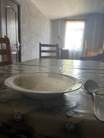 A white bowl of soup is on a table covered with a patterned plastic tablecloth. A silver spoon lies to the right of the bowl. In the background, a wooden chair is visible, and further back, a doorway with a door is on the left. Sunlight streams through a window with sheer curtains on the right. The environment appears to be an interior space, possibly a dining area in a dwelling within the ღრმაღელის თემი region of Georgia. The overall lighting is bright, with some shadows.
