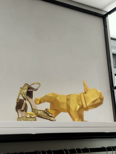 This indoor image captures a stylish display, likely within a retail setting, in Bellinzona, Switzerland. The central subjects are a pair of glittering gold high-heeled sandals and a golden, polygonal sculpture of a French bulldog, both resting on a clean white shelf.
The sandals are a prominent feature, covered in gold glitter and adorned with large, square-cut yellow gemstone-like embellishments over the toe straps. The heels are slim and golden, and ankle straps are visible. Partial text can be seen on the inner lining of one shoe, reading "CASPERI", and "ERIN" is visible on another part of the shoe.
Beside the shoes, the golden French bulldog sculpture is rendered in a distinct geometric, low-poly style, composed of many flat facets that reflect light. It stands in a dynamic, slightly forward-leaning pose.
The background is a plain, light gray wall, creating a minimalist backdrop that allows the golden items to stand out. A black metal frame is visible along the top and right edge of the image, suggesting the structure of a display cabinet or shelving unit. A darker, possibly metallic, bar runs horizontally below the white shelf, further indicating a display setup. The lighting is bright and even, typical of an indoor retail environment. No human activity is depicted, emphasizing the objects themselves.