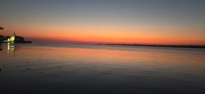 The image depicts a serene sunset over a body of water, likely a lake, with a distant shoreline. The sky transitions from a soft blue at the top to vibrant oranges and reds near the horizon, indicating it is dusk. The water reflects the colors of the sky, creating a warm and peaceful ambiance.

On the left side of the frame, there are structures that appear to be part of a waterfront development, possibly including a building with a distinctive sail-like roof illuminated by artificial lights. A dock or pier extends into the water from this area. A few dark objects, possibly buoys or debris, are visible on the water's surface closer to the foreground. The distant shoreline is a dark silhouette against the colorful sky, with hints of lights suggesting human habitation.

The overall mood is tranquil and contemplative, evoking a sense of calm at the end of the day. The weather appears to be clear, allowing for a dramatic and beautiful sunset. The location context of Bregenz, Austria, suggests this could be a view of the Bodensee (Lake Constance). There is no visible text in the image.
