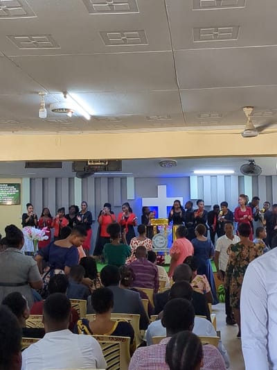 The image shows an indoor gathering taking place in a church in Morogoro, Tanzania (coordinates: -6.824, 37.677). The room has a tiled floor and a ceiling with rectangular panels. A group of people are seated in chairs facing a stage. On the stage, a group of people are singing. They are wearing various colored clothing and are holding microphones. There is a white cross on the stage behind the singers. The image is captured from the perspective of someone seated in the audience, looking towards the stage. The people in the audience are seated in rows, and some are engaged in conversations. The image captures a moment of worship and community in a church setting.