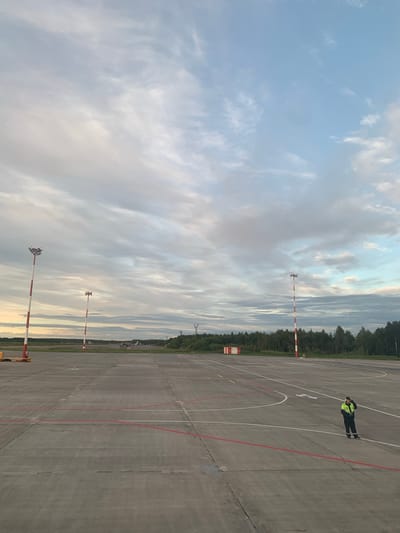 The image depicts an airfield with a concrete tarmac marked with red and white lines. Several tall light poles, striped red and white, are visible on the tarmac. In the distance, a line of trees forms the horizon, with a few buildings and aircraft interspersed. The sky is partly cloudy with hues of blue, grey, and white. On the tarmac, a person wearing a reflective yellow vest and dark pants is standing. This scene is located at the Arkhangelsk Airport (Talagi Airport) in Arkhangelsk, Russia.
