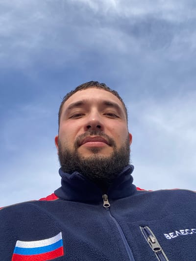 A man with a dark beard, wearing a navy blue fleece jacket with a Russian flag patch on the chest and "BEAECC" embroidered on the upper right, is posing for a selfie under a bright, partly cloudy sky. The shot is taken from a low angle, looking up at the man. The location is possibly Koskolovo, Russia, given the flag patch.