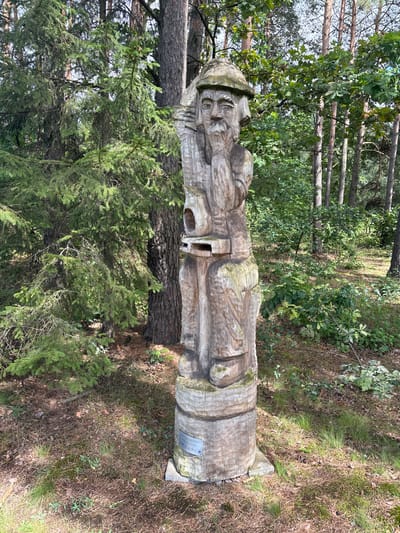 The image shows a wooden sculpture of a man's head and upper body, carved in a folk art style. The sculpture is weathered and stands on a cylindrical base. It is positioned outdoors in a forest or park setting, with trees and foliage visible in the background. A small plaque is attached to the base of the sculpture. The location is identified as Sielachowskie, Poland.