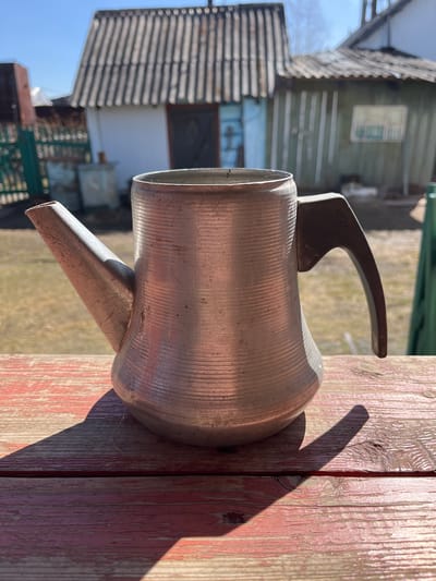 An aluminum watering can is positioned on a weathered wooden surface with red paint in Верхняя Уря, Russia. The can is oriented so that the spout extends downward. The handle is curved and attached to the top of the can. The surface on which the can rests shows distinct linear patterns and texture of wood. In the background, elements of a rural setting are visible, including buildings with shingled roofs and weathered siding. Sunlight casts shadows across the objects and the wooden surface.