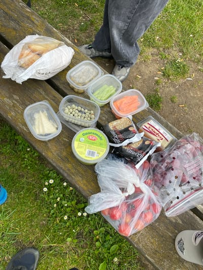 On a weathered wooden bench outdoors in Villaviciosa, Spain, a picnic is laid out. The scene shows food items including:
* Grapes and tomatoes in plastic bags
* A container of guacamole
* Rice cakes
* A block of cheese
* Several clear plastic containers holding carrots, celery, olives, capers, and pieces of cheese.
* A white plastic bag filled with bread.
A water bottle is visible towards the top of the image. The ground nearby is covered in grass, with cigarette butts and small white flowers scattered around. A person's leg wearing jeans and grey sneakers is visible to the left. Another person wearing a shoe can be seen on the bottom right of the image.