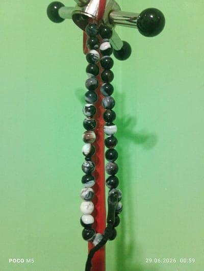 A vertical, close-up shot shows a string of dark and light-patterned beads hanging against a pale green wall. The beads appear to be made of polished stone and are spherical in shape, with a variety of marbled patterns of black, white, and gray. The string is attached to a metal apparatus with black spherical ends, suggesting it might be a part of a prayer bead set or a decorative item. The bottom of the string shows a black tassel or a knot. In the lower left corner, the text "POCO M5" is visible, and in the lower right corner, the date and time "29.06.2026 00:59" are displayed. The lighting creates a soft shadow of the beads on the green wall. There are no people or animals in the image, and the scene is static. The image was taken indoors, likely in a residential setting. There is no information available to suggest this image is located in the city of Hisor, Tajikistan.