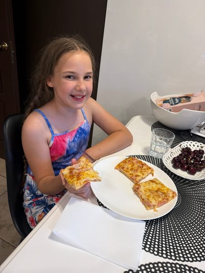 In Baloži, Latvia, a young girl sits at a white table, holding a slice of pizza. She is wearing a colorful tie-dye dress. In front of her are three more slices of pizza on a white plate, a glass of water, a bowl of cherries, and a white napkin. The background shows a dark door and a partially visible shelf with boxes of ice cream. The scene appears to be indoors, likely a dining area.