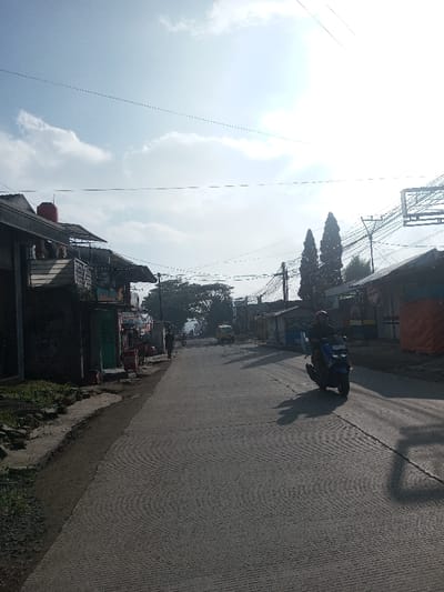 A long, straight road leads into the distance, flanked by modest buildings on either side. Utility poles and wires crisscross overhead. A person rides a scooter toward the viewer on the right side of the road, casting a long shadow. Another person walks along the left side of the street. The sky is bright with scattered clouds. Two tall, slender trees stand in the distance to the right.