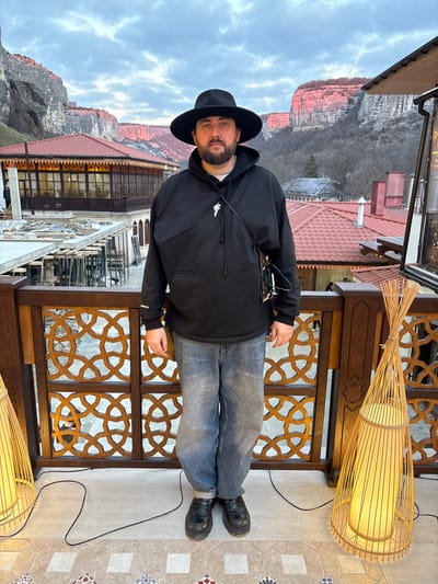 A male individual stands centered on an outdoor tiled patio or balcony, facing directly towards the viewer. He wears a black wide-brimmed hat, an oversized black hooded sweatshirt with a white stylized symbol on the left chest, faded blue denim jeans, and black chunky-soled slip-on shoes. A thin black strap crosses his chest, from which dark-rimmed glasses hang. He has a dark beard and visible dark hair.

The patio floor features a repeating geometric tile pattern in light gray, white, red-brown, and dark blue, with a dark electrical cord running across it. A waist-high wooden railing with intricate geometric latticework separates the patio from the background. Two tall, conical floor lamps with bamboo-like slats and internal diffusers, emitting a warm yellow light, flank the individual.

In the midground, beyond the railing, multiple buildings with red-tiled roofs are visible. To the left, a multi-story building with large windows is under apparent construction, indicated by scaffolding. Other buildings with traditional architectural elements are scattered across the scene.

The background is dominated by large, steep rock formations or cliffs, characteristic of a mountainous region. The cliffs on the right exhibit a distinct reddish-orange glow, suggesting direct illumination from a low sun (sunrise or sunset). The sky above is overcast with diffused light, displaying shades of light blue and gray. The architectural styles, red-tiled roofs, and prominent cliff formations are consistent with the city of Bakhchysarai, Ukraine, specifically within a valley or elevated area providing scenic views of the surrounding Crimean Mountains.