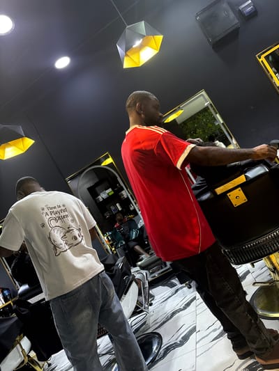 The image depicts two men in what appears to be a barbershop or salon in Lugbe, Nigeria. One man, wearing a red shirt with white stripes, is seated in a barber's chair. The second man, wearing a white t-shirt with a cat graphic, is standing next to the chair, possibly a barber. The floor is tiled with a black and white marble pattern. There is a mirror in the background, reflecting the shop's interior. The lighting is modern, featuring geometric light fixtures. Shelves holding various products are visible.