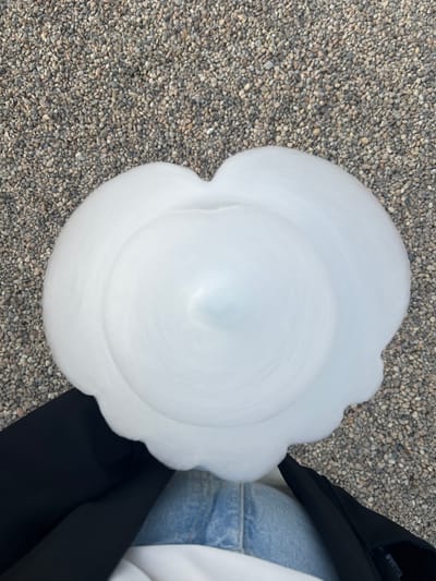 This is a top-down, medium shot of a large, fluffy, white mass of what appears to be cotton candy, or a similar spun sugar confection, resting on a gravel surface. The cotton candy is somewhat amorphous, with soft, undulating edges, and a swirled, conical shape in the center. A person's legs are visible at the bottom of the frame; they are wearing blue jeans and a dark jacket. The background consists of small, gray and brown pebbles. The scene appears to be outdoors, on a pathway or ground cover composed of gravel. No specific landmarks or identifiable buildings are visible that would definitively place the location in Listvyanka, Russia.