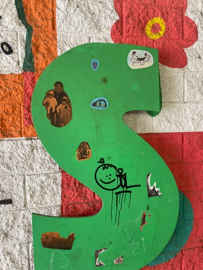 The image shows a large, green, S-shaped cutout positioned against a textured white brick wall. The cutout is adorned with several small stickers and some drawn elements. At the top of the S, a sticker depicts a simple drawing of two stick figures interacting at a table. Below this, another sticker shows a small blue camper van. To the left, a collage of photos shows a woman holding two young children. Further down the S, a sticker displays a figure riding a motorcycle, and another sticker contains an image of an urban landscape. A black line drawing of a smiling face with arms and legs is also present on the green surface, with black drips extending from it. A crudely drawn heart and the number "21" are visible towards the bottom. Various other smaller, unidentifiable stickers and markings are scattered across the green cutout. The background wall is painted with large, abstract shapes in orange, red, and green. The overall scene suggests an outdoor public art installation or graffiti.

This artwork is located on the wall of the gymnasium of the Escola de Futbol Fundació Badalona, in Badalona, Spain. The artwork is part of a mural project titled "Art per l'Esport" (Art for Sport).