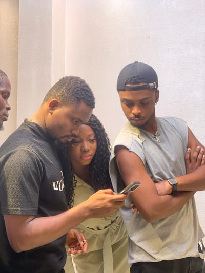 An indoor scene in Moba, Nigeria, depicts four individuals of African descent gathered around a smartphone. The background consists of textured, light-colored walls. In the central foreground, a male with short dark hair and a beard, wearing a black textured short-sleeved shirt, holds a silver smartphone in his left hand, with his gaze directed at its screen. To his immediate right, a female with long dark wavy hair, dressed in a light-colored collared garment with a tie-waist, also looks intently at the phone. To the far left, a partially visible male figure with short dark hair is oriented towards the phone. To the right, a male wearing a backward black baseball cap, a grey ripped sleeveless shirt, a silver chain with a "4K" pendant, and a dark-strapped watch on his left wrist, stands with his arms crossed, his head tilted downwards towards the phone screen. All visible individuals are focused on the smartphone's display.