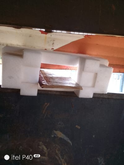 The image depicts a confined space, possibly an interior of a structure, in Hoche, Nigeria. The upper portion of the frame shows a dark, likely metallic, horizontal surface. Below this, a white structural beam, exhibiting rust and red paint residue, runs horizontally, with a visible pattern of small perforations. Behind the beam and to its right, an orange surface is partially visible. Centrally, a piece of white, molded polystyrene packaging material is positioned. Within the polystyrene's cavity rests a stack of items, appearing to be flat, possibly books or documents, wrapped in clear plastic and featuring a reddish-brown, patterned cover. A light-colored, possibly wooden or cardboard, base supports these items. The lower section of the frame consists of a dark, textured, and possibly worn surface. Natural light enters from the right side, illuminating the central area. No individuals or actions are depicted. The scene suggests an industrial or storage environment.