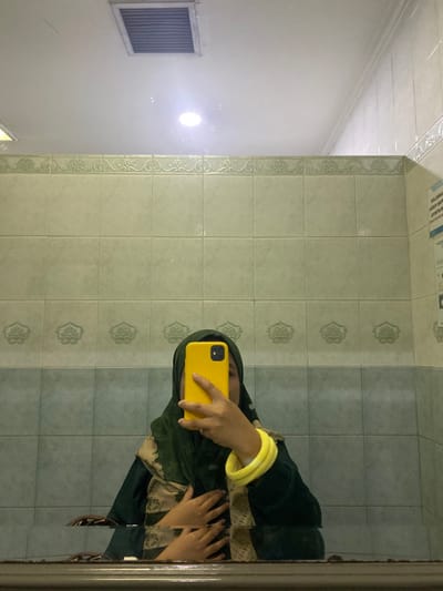 The image depicts a mirror selfie taken by an individual in an indoor tiled room, consistent with a public or commercial restroom or waiting area, located in Cipeundeuy, Indonesia.
An individual, wearing a dark green hijab and a dark green garment with a beige patterned section on the front, is visible from the chest up. Their face is entirely obscured by a bright yellow smartphone, held in their left hand in a selfie orientation. The individual's right hand is placed over their chest area, and two bright yellow bangles are worn on their right wrist.
The background consists of tiled walls and a ceiling. The lower portion of the wall features light blue-green square tiles. Above this, a decorative border of light green tiles with a repeating scrollwork pattern runs horizontally. The upper portion of the wall is composed of light green square tiles. On the right side of the image, a partially visible white rectangular sign or poster is present on the tiled wall, displaying mirrored Indonesian text. The white ceiling above the tiles features a rectangular ventilation grate and an illuminated circular recessed light fixture, casting a bright reflection in the mirror. The lower edge of the mirror frame or a counter is visible at the bottom of the image.