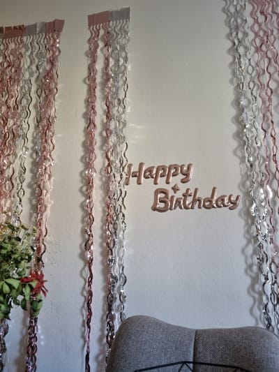 This image shows a close-up, angled view of a white wall decorated with metallic streamers and a "Happy Birthday" balloon. The streamers are arranged in horizontal rows, alternating between pink and silver. They are wavy and appear to be made of thin, reflective material.
The "Happy Birthday" balloon is a rose gold color and is positioned vertically on the wall, with "Happy" above "Birthday". There is also a small, star-shaped balloon or decoration next to "Birthday".
To the right of the balloon, a portion of a gray, textured object is visible, possibly a mannequin or piece of furniture with fabric covering. In the bottom right corner, a small arrangement of dried flowers and greenery is partially visible, with a single red flower or leaf.
The lighting suggests an indoor setting, with shadows cast by the streamers and balloon on the wall, indicating the light source is from above and slightly to the left. The overall impression is of a celebratory decoration for a birthday party. The scene appears to be set in Saarbrücken, Germany.