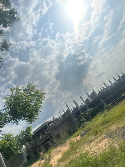 This image depicts an unfinished, multi-story concrete structure in Amansee, Nigeria. The building appears to be in a state of construction, with exposed rebar protruding from the upper levels. The structure is surrounded by lush green vegetation and a sandy, uneven terrain. The sky is filled with a mix of white and gray clouds, with the sun visible in the upper left portion of the frame, suggesting it is daytime. The perspective of the image is unusual, tilted at an angle, making it appear as if the building and the landscape are leaning. There are no people visible in the image, but the presence of the building suggests ongoing construction or a paused development project. The overall impression is one of an incomplete structure in a natural, somewhat overgrown environment. The weather appears to be fair with scattered clouds.