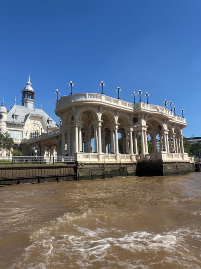 The image shows the Tigre Art Museum, formerly the Tigre Club, in Tigre, Buenos Aires, Argentina, from a low angle, likely taken from a boat or near the water's edge. The museum features a multi-story structure with an ornate architectural style, including columns, arches, and decorative elements. Small spherical lamps line the top of the building. The building extends over the brown, moving water, which forms the right side of the photo. The sky is a clear, bright blue.