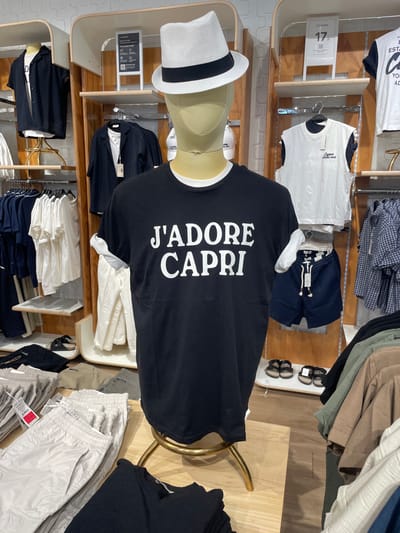 The image shows a retail display of men's clothing in a store, likely located in Marcianise, Italy, as indicated by the context of a previous query. In the foreground, a mannequin wearing a black t-shirt with white lettering that reads "J'ADORE CAPRI" is displayed. The mannequin is wearing a white fedora hat with a black band. Behind the mannequin, various clothing items are neatly arranged on shelves and hanging racks. These include shirts, shorts, and what appear to be pants or tracksuits in neutral colors like white, beige, and olive green. A price tag of "17.99" is visible on a white t-shirt displayed on a shelf. A pair of sandals is also visible on a lower shelf. The overall environment suggests a well-organized clothing store display.