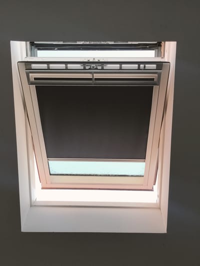 The image shows an interior view looking up at an open skylight. The skylight has a white frame and a dark roller blind partially covering it. The window pane itself appears to be dirty. The interior walls are painted a dark gray color. The skylight appears to be a standard residential type, possibly a Velux brand, judging by the faint text visible on the frame. There is no indication of the location being in Perpignan, France; the image is a generic interior shot of a skylight.