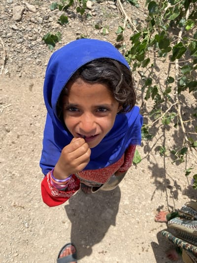 A young girl in Ashoqah, Afghanistan, is depicted in a high-angle, close-up portrait, looking upwards directly at the camera with a gentle, curious expression. She wears a bright blue headscarf that covers her hair, with some dark, curly strands visible around her face, secured by a small silver clip. Her dark eyes are bright and engaging. She is dressed in a red top with a white or light-colored pattern, and multiple colorful bangles adorn her left wrist. Her right hand is raised near her mouth, suggesting she might be holding something small or performing a natural gesture.
The scene is set outdoors on what appears to be dry, light-colored dirt or gravel ground, strewn with small stones. In the background and extending along the right side of the frame, there are green leaves and branches of a plant or tree, casting distinct, dappled shadows on the ground. These strong shadows indicate bright, direct sunlight, suggesting the time of day is likely midday or early afternoon with clear weather. In the lower right corner, the bare feet of another person wearing a long, patterned skirt or dress are visible. The bare foot of a third person, wearing a grey sandal, is partially seen in the lower left corner. There is no visible text in the image. The overall impression is a candid moment of interaction in a sunlit, rural environment.