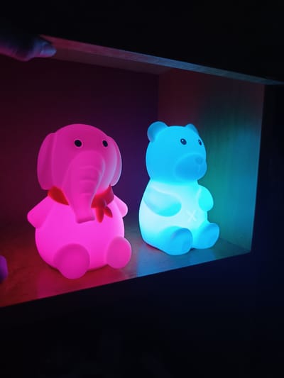 Two illuminated, animal-shaped night lights sit on a shelf within a dark, enclosed space. The left figure is an elephant, lit in bright pink, with a red ribbon around its neck. The right figure is a teddy bear, lit in bright blue, with a white "X" on its chest. The top of a finger is visible in the upper left corner, near the shelf edge. The location, Cherkovitsa, Bulgaria, is irrelevant as there is no visible information in the image to confirm this.