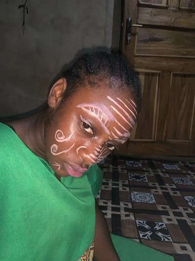 A medium shot captures a young Black person adorned with white face paint, looking downwards. The face paint features parallel lines across the forehead and temples, swirling patterns on the cheeks, and a small V-shape below the lip. The individual wears a bright green garment and is positioned against a dark, textured wall. To the right, a wooden door with horizontal panels is visible. The foreground shows a patterned surface with brown, black, and white squares. The overall lighting is dim, suggesting an indoor setting. The location is identified as Agban, Nigeria.