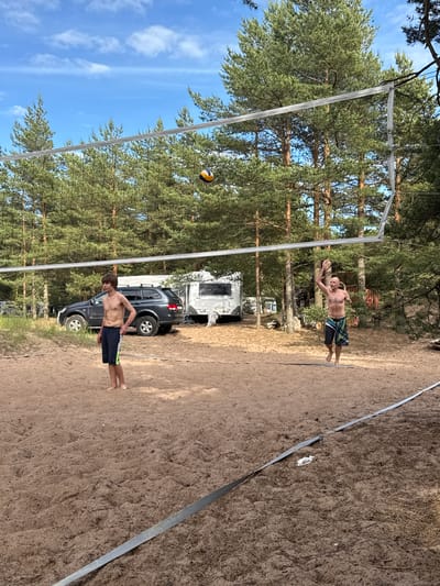 In Zelenaya Roshcha, Russia, two shirtless men are playing volleyball on a sand court surrounded by pine trees. A volleyball is in mid-air, above the net. In the background, a dark SUV and a camper trailer are parked near the trees. The sky is blue with scattered white clouds. The man on the left is wearing dark shorts with a green stripe and is standing facing forward. The man on the right is wearing patterned shorts and is in the process of jumping with his arm raised. The court has white boundary lines.