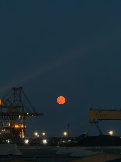 The image depicts a dusk or nighttime scene at the Port of Constanța, Romania. A large, bright orange moon hangs in the dark blue sky. In the foreground and mid-ground, industrial structures are visible. To the left, a large yellow gantry crane is illuminated by orange lights. To the right, another yellow structure, possibly a conveyor system or part of a crane, is also visible. Several streetlights and other artificial lights are scattered throughout the scene, illuminating industrial elements and pathways. In the distance, dark silhouettes of hills or mounds can be seen against the sky. The overall impression is of a working port environment under the glow of the moon and artificial lighting.