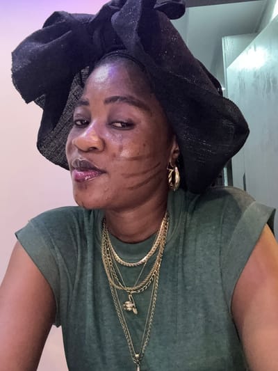 The media file depicts a close-up, head-and-shoulders shot of a woman in Ajara, Nigeria. She is wearing a black headwrap and a green t-shirt. Multiple gold necklaces are layered around her neck. She has dark skin and appears to be wearing makeup, including lip gloss and what looks like body paint or lines drawn on her cheeks. Her expression is directed towards the camera, with her eyes open and lips slightly parted. The lighting suggests an indoor setting, possibly artificial light, with a hint of purple light on the left side of the frame. The background is blurred, with indistinct shapes and textures, including what appears to be a reflective surface above and to the left of the subject. There is no visible text in the image. The overall mood conveyed is one of self-presentation or a casual portrait.