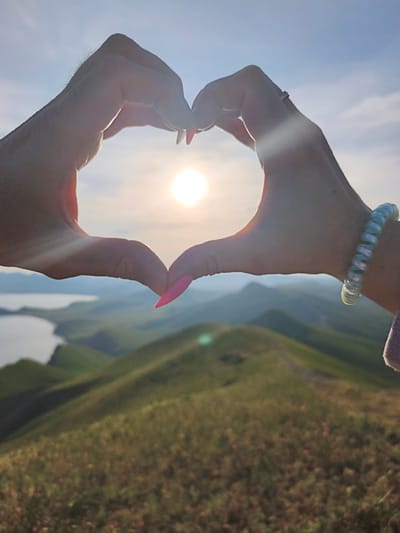 Two hands form a heart shape against a backdrop of rolling hills and a body of water, likely the Black Sea. The sun is positioned centrally within the heart, creating a glowing effect. The hills are covered in vegetation. The hand on the right has a spiral hair tie around the wrist and a ring on the finger. The hand on the left has pink fingernails. The location is likely near Коктебельский поселковый совет, Ukraine, judging by the landscape.