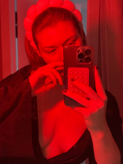 This image is a mirror selfie taken indoors in Nikol'skii, Russia, under strong red lighting. The central subject is an individual with hair pulled back, wearing a light-colored, segmented headband and a dark, open top or robe that reveals a portion of their upper chest. A dark strap with white markings is visible on the left side of the chest area. The subject's right hand holds a dark-colored smartphone with a glittery case and a triple-lens camera module. Attached to the back of the phone is a red, rectangular accessory featuring a grid of circular indentations. The subject's left hand is positioned near their lower face. Fingernails on both visible hands are painted dark. The individual is looking downwards, likely at the phone screen. The background is largely obscured by the red light, showing only faint reflections and a vertical line suggesting an architectural element like a doorframe.