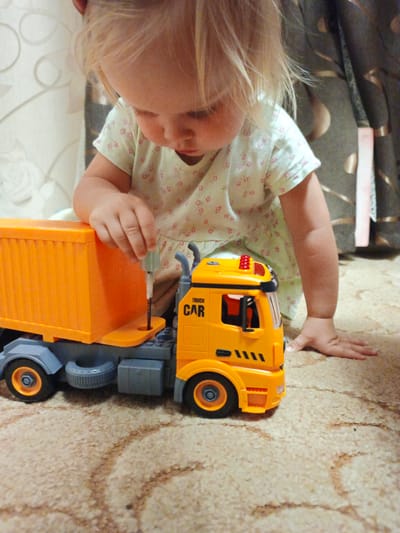 A blonde toddler with light skin, wearing a pale green t-shirt with a pink floral pattern, is kneeling on a beige carpeted floor. The child is looking down intently at a toy truck. The truck is orange and grey, with a detachable orange trailer. The child's right hand is reaching out, holding a small, silver screwdriver, with the tip inserted into a hole on the truck's chassis. The environment appears to be an indoor setting, possibly a living room, with a curtain visible in the background. The image is taken from a slightly elevated angle, focusing on the child and the toy. This scene is unlikely to be specifically located in Zlatoust, Russia, as there are no identifying geographical or architectural markers.