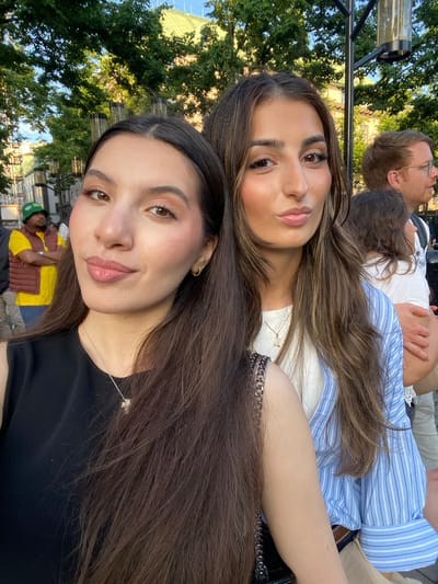 Two young women are taking a selfie outdoors in what appears to be a city environment. The woman on the left is wearing a black sleeveless top and a silver necklace. The woman on the right is wearing a white top layered under a blue and white striped button-down shirt. They are standing close together, with the woman on the right's arm resting on the woman on the left's shoulder. In the background, other people are visible, including a man in a yellow shirt and a green hat, and a man with glasses. Trees and buildings can be seen behind them, suggesting an urban park or a street. The lighting indicates it is daytime, likely late afternoon given the warm tones. Based on the architectural style of the buildings and the presence of a street lamp consistent with the city's urban design, the scene is likely in Stockholm, Sweden.