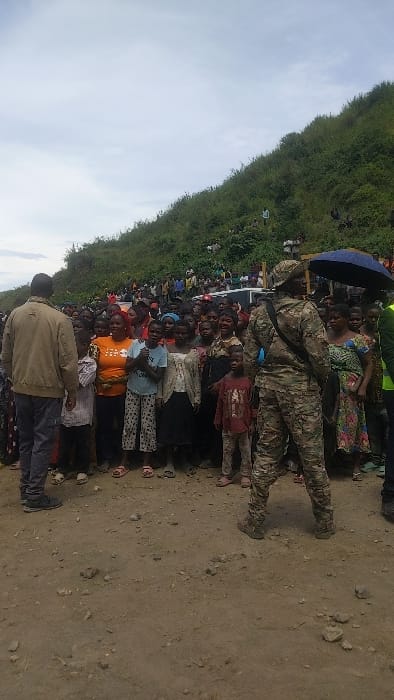 The image shows a scene from Sake, Democratic Republic of Congo, at coordinates -1.57 latitude and 29.046 longitude. It depicts a large gathering of people in front of a densely vegetated hillside. A man wearing a brown jacket and grey pants stands at the left edge of the frame, looking towards the center. A group of people, including women and children, are arranged in front of him. Some of the women are holding babies. A man wearing a camouflage military uniform and a black strap across his chest stands at the right edge of the frame, facing the group. He is holding a blue umbrella that is partially obscuring the group behind him. There is a white vehicle parked in the background behind the group of people. The ground is dirt and covered in loose rocks and pebbles. The sky is overcast and a hill covered in green vegetation dominates the background. A large number of people are on the hillside, out of focus, suggesting a large gathering. The scene seems to be taking place in a rural area, possibly a village or an open-air market.