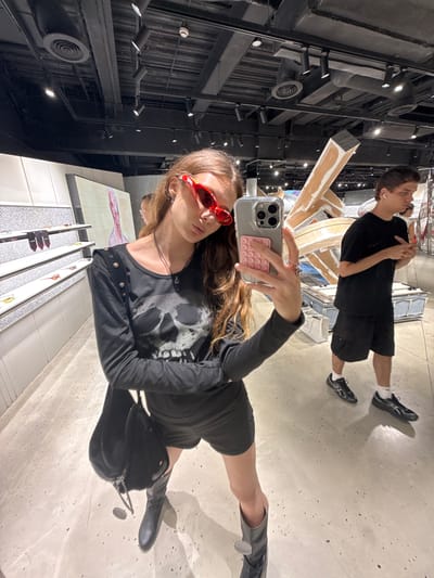 A young woman is featured prominently in the foreground, taking a mirror selfie in an indoor setting. She has long brown hair and is wearing distinctive red cat-eye sunglasses. Her attire includes a black long-sleeved top with a large white skull graphic, a black skirt, and black boots. She holds a grey smartphone, fitted with a transparent case and a pink pop-it accessory, directly towards the reflective surface. Her expression is posed and direct.
In the background, a young man stands further away, reflected in the same mirror. He has short brown hair and is dressed in a black t-shirt, black shorts, and black athletic shoes, with white wireless earbuds in his ears. His posture is relaxed.
The scene appears to be a modern commercial space, likely a retail store or gallery, located in Shanghai, China. The floor is light-colored and somewhat reflective, contributing to the selfie effect. The ceiling is dark and industrial-style, featuring exposed pipes, ventilation systems, and multiple track lighting fixtures. Various display structures and architectural elements are partially visible around the subjects. The overall aesthetic is contemporary and urban. No text is explicitly visible in the image.