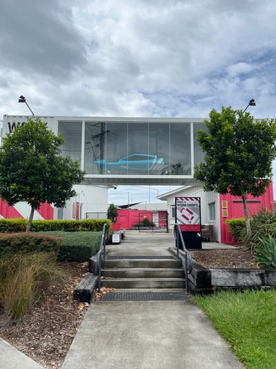 This image depicts an outdoor commercial or industrial area in Hamilton, Australia, characterized by repurposed shipping container architecture under an overcast sky.

In the foreground and mid-ground, a concrete pathway leads past landscaped sections featuring green grass, shrubs, mulch, and several small trees. To the right of the path, a set of concrete steps with metal railings ascends to a higher level.

On the left side of the scene, a multi-story building combines white shipping containers at its base with a large, two-story glass facade above. Displayed prominently within the upper glass section is a large, light blue object with white stripes, resembling a marine vessel or boat. Visible text on this object reads "NORTH SHORE". A black streetlight is mounted near the top of this building.

Further into the scene, another sign stands prominently beside the path, featuring the text "DOCK C WORKSHOPS" and an illustrative graphic, possibly depicting an interior space or furniture. Beyond this, several other buildings constructed from red and white shipping containers are visible, contributing to the distinct aesthetic of the location.

The scene appears quiet, with no people visible. The time of day is daytime, under cloudy conditions.