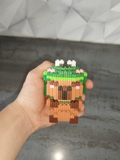 A hand is holding a small, multi-colored toy figure made of interlocking plastic bricks. The figure appears to be a stylized bear with a green, leafy headpiece that has white and pink accents. The bear's body is brown with darker brown and pink details, representing features like its nose, ears, and paws. The background consists of a textured grey surface below and a patterned white surface above, possibly a wall and a countertop or floor. The image's lighting suggests it was taken indoors, likely in a home environment. There is no indication of the city of Severodvinsk, Russia, or any specific urban location within the image or its provided crops.