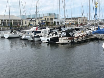The image shows a marina in Wismar, Germany, on a cloudy day. Several sailboats are docked in the water, with their masts reaching towards the sky. The boats are of various sizes and designs, with some featuring white hulls and others with darker colors. In the background, there are industrial buildings and cranes, suggesting that this is a working port as well as a recreational area. The water is calm, with gentle ripples reflecting the overcast sky. There are no people visible in the image, and the scene is peaceful and still. The overall impression is one of a quiet, functional harbor, typical of a coastal city.