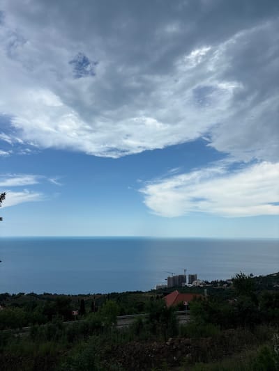 The image depicts a scenic view of the Black Sea coast near Malyi Mayak, Ukraine. The horizon is dominated by the vast expanse of the deep blue sea meeting a sky filled with a dramatic mix of white and gray clouds. In the mid-ground, a lush green landscape rolls down towards the sea, dotted with trees and vegetation. Nestled within this greenery are buildings, including some under construction with cranes visible, suggesting development in the area. A road runs through the foreground, bordered by dense foliage and dark soil. Road signs are present on the side of the road, indicating traffic direction. The overall impression is one of natural beauty contrasted with ongoing human activity.
