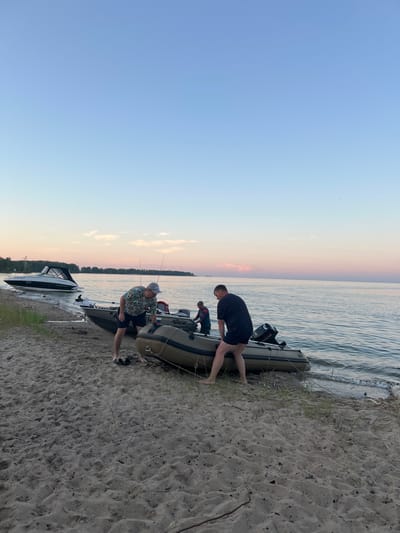 The image depicts a sandy beach along a body of water, likely a lake or sea, under a pale blue sky with hints of pink and orange from a sunset. In the foreground, three men are interacting with two inflatable boats and one rigid-hulled boat. Two of the men are actively engaged with an inflatable boat that is partially submerged in the water, appearing to be either launching or retrieving it. One man, wearing a patterned shirt and shorts, is crouched down next to the boat, while another, in a black t-shirt and shorts, is pushing the boat from the water. A third, younger man, is positioned between the two boats, partially obscured. The rigid-hulled boat is further up on the sand, with its bow pointed towards the water. A larger, white boat with a cabin is anchored offshore in the background, with a tree-lined shore visible in the distance. The scene suggests a recreational activity, possibly fishing or boating, taking place at dusk. The location is identified as the city of Borovoye, Russia.