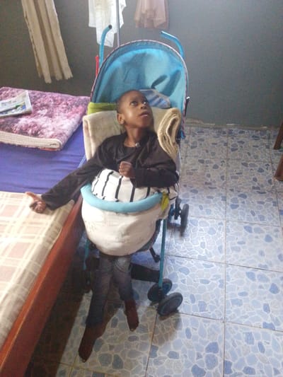In Lumakanda ward, Kenya, a child sits in a blue and silver stroller. The child is wearing a dark long-sleeved shirt and striped pants. The stroller is placed next to a bed with a pink and white patterned blanket. The floor is tiled with a blue and white mosaic pattern. Curtains hang in the background. The image appears to be taken indoors.