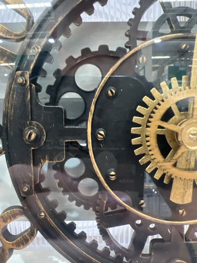 The image is a close-up, eye-level, medium shot of a decorative clock mechanism. The clock is made of metal with an antique, distressed gold and black finish. Gears of various sizes are interlocked and visible through a clear glass casing. Some gears are large and dark, while others are smaller and a brighter gold color. The gears are intricate and have visible teeth. There are screws holding some of the components together. In the background, through the glass and the spaces between the gears, a blurry, light-colored brick pattern is visible. The lighting is somewhat diffuse, and there are reflections on the glass surface. No people are visible. The focus is entirely on the detailed clockwork. There is no visible text in the image. The scene appears to be indoors, possibly a home or shop, given the decorative nature of the clock. The time of day and weather are not discernible.