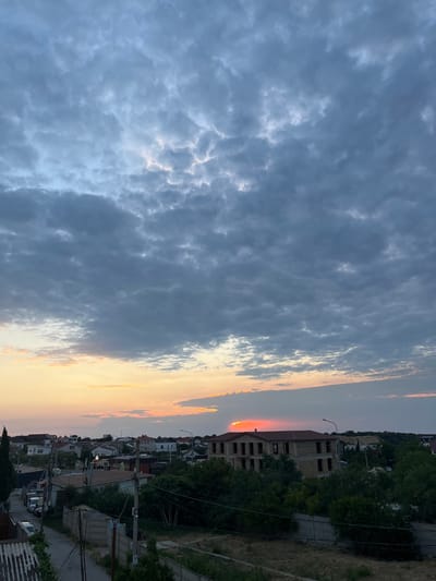 The image depicts a sunset over a residential area in Balaklavsky District, Ukraine. The sky is filled with dramatic, dark gray clouds, interspersed with streaks of orange and pink from the setting sun. A vibrant orange glow emanates from behind a large, unfinished brick building in the mid-ground. This building appears to be under construction, with exposed window frames and a dark roof.
The foreground shows a dirt road or pathway with a concrete fence on the right, and what appears to be a wooden fence or corrugated metal structure on the left. Several trees and dense green foliage surround the buildings, creating a lush, natural element within the urban landscape. In the distance, a line of houses with red and white roofs is visible, interspersed with trees and utility poles. Parked cars are lining the street on the left side of the image. The overall impression is one of a quiet, developing residential area experiencing a beautiful sunset.