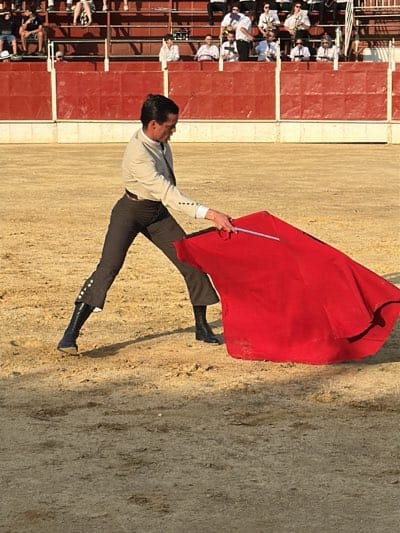 The image depicts a bullfighter in a sandy arena in San José del Valle, Spain. He is wearing dark pants with buttoned detailing at the lower leg, black boots, and a light-colored shirt. He holds a large, red cape in his left hand, extending it away from his body. A wooden support stick is visible within the cape. Spectators are seated in tiered seating in the background, behind a red barrier. The lighting suggests a sunny day.
