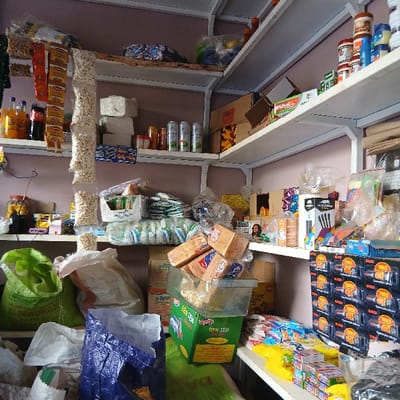 The image displays the interior of a retail general store in Moshi, Tanzania. The space is characterized by white metal-frame shelving units heavily laden with merchandise, extending along the visible walls.
On the upper left, multiple clear plastic bags of popcorn hang vertically alongside strips of orange-packaged snacks. Below these, various beverages, including Coca-Cola and Fanta, are arranged with stacks of white paper products and green-labeled aerosol cans.
The upper right shelves feature an assortment of packaged food items, including jars, cans, and cardboard boxes. An open cardboard box is also visible.
Mid-level and lower sections contain large green and white polypropylene sacks, appearing to hold bulk grains or pulses. One white sack has handwritten markings. A clear plastic container, filled with popcorn, rests on a green box labeled "BEN TEN." Stacked loaves of bread in clear plastic wrappers are positioned on top of the popcorn container.
The lower right shelving and counter display numerous small packaged goods, such as biscuits, candies, and 'POWER' branded boxes, likely containing batteries or small electronics. A crumpled blue plastic bag lies on the lower left.
The walls are painted a pale pink or purple hue, and the ceiling is white. No individuals are present, and no actions or interactions are depicted within the static scene.